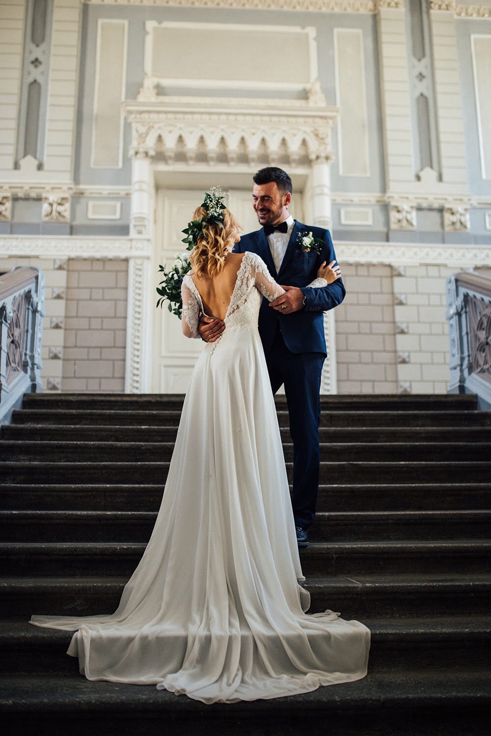 Para młoda na schodach w eleganckim budynku, panna młoda w sukni z koronkowymi rękawami i wiankiem, pan młody w granatowym garniturze z muszką, uśmiechnięci i obejmujący się.