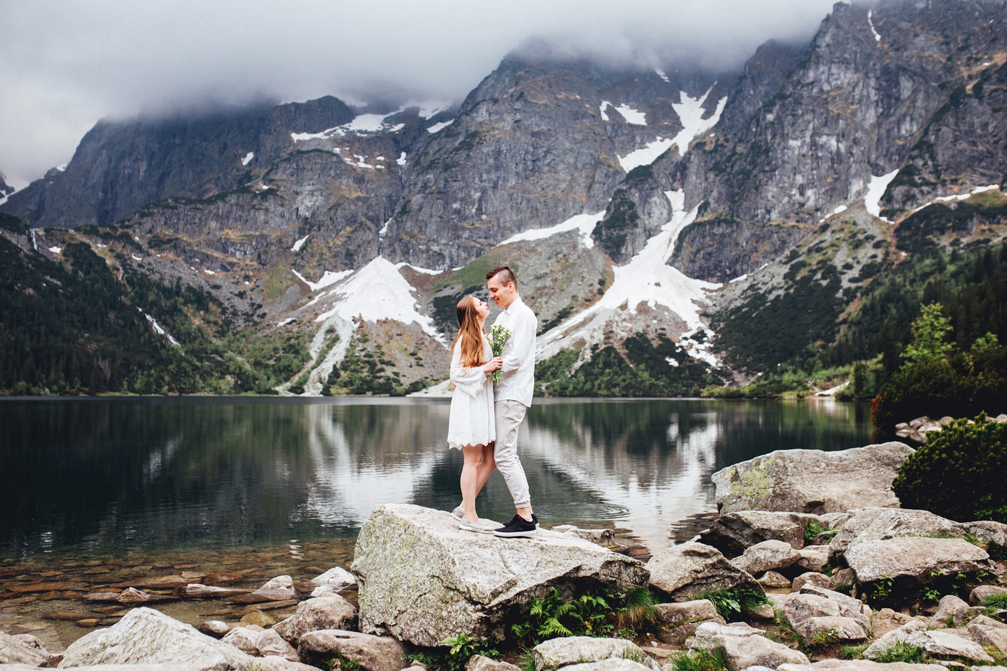 Para młoda w białych strojach stoi na skale nad brzegiem górskiego jeziora, w tle szczyty górskie spowite mgłą.
