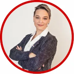 Portret uśmiechniętej kobiety w marynarce z przypinką agencji nieruchomości, ręce skrzyżowane, na jednolitym tle w czerwonej ramce.