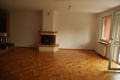 Jasny pokój z parkietem ułożonym w jodełkę, kominkiem z ceglaną obudową i otwartymi drzwiami balkonowymi w słoneczny dzień.