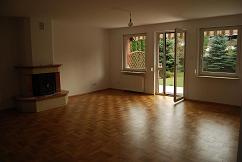 Pusty pokój z parkietem ułożonym we wzór jodełki, kominkiem z ceglaną obudową i otwartymi drzwiami balkonowymi wychodzącymi na ogród.