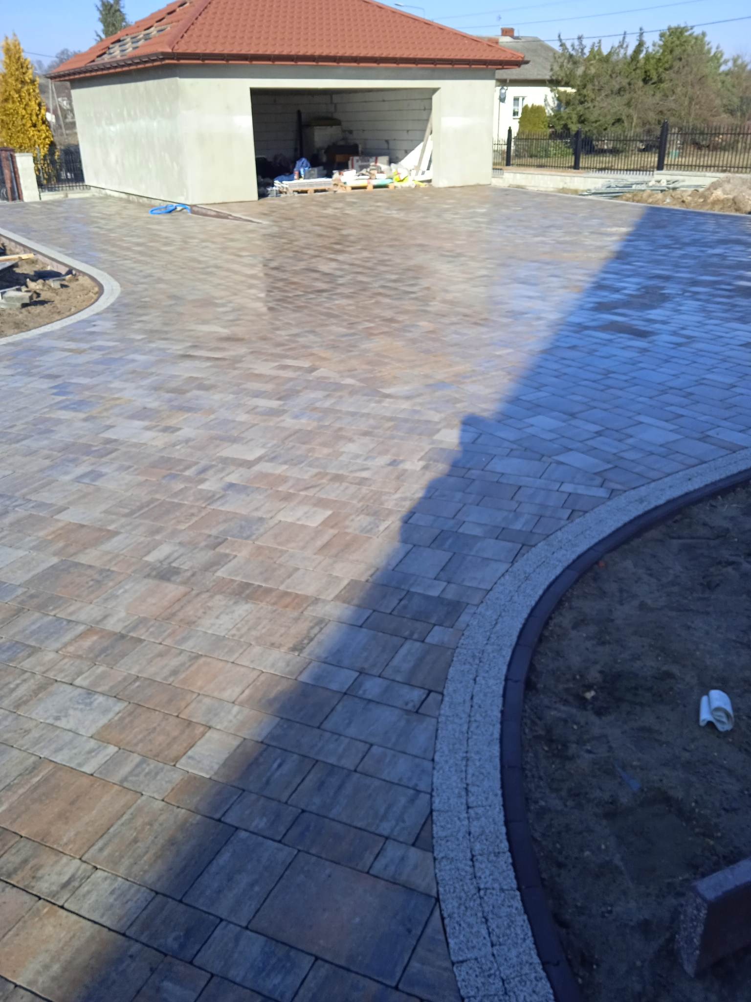 Nowo ułożona, wielokolorowa kostka brukowa na podjeździe przed domem z garażem, obrzeża wykończone kostką granitową, widoczne ślady po pracy i materiały budowlane w garażu.