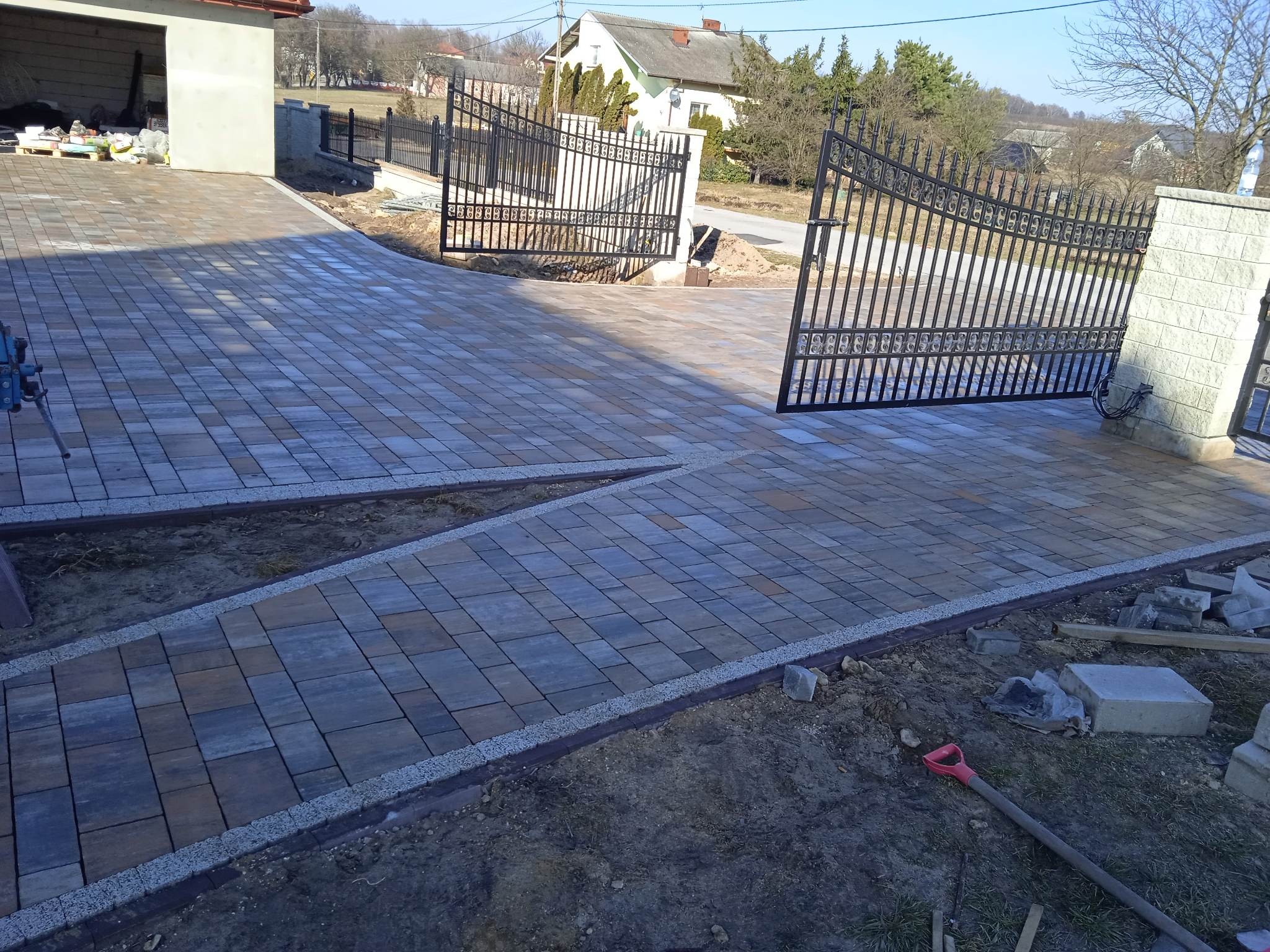 Ułożona kostka brukowa prowadząca do bramy wjazdowej z elementami niedokończonej pracy: widoczna łopata, kostki i ziemia.