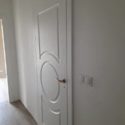 Białe drzwi wewnętrzne z okrągłym motywem dekoracyjnym i złotą klamką, zamontowane w jasnym pomieszczeniu z widocznym fragmentem podłogi i włącznikiem światła.