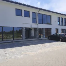 Nowoczesny budynek biurowy z dużymi, czarnymi oknami, jasną elewacją i kamiennym wykończeniem przy wejściu, zaparkowany czarny samoch&oacute;d osobowy przed budynkiem.