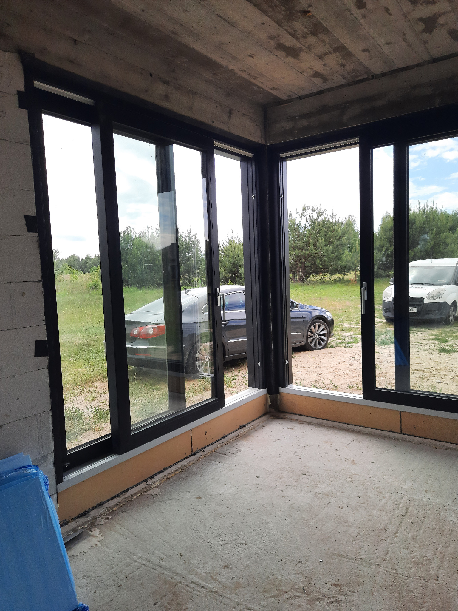 Surowy stan pomieszczenia z dużymi oknami przesuwnymi z czarnymi ramami; widok na zewnątrz z samochodami na trawie, betonowy sufit i podłoga w trakcie wykończenia.