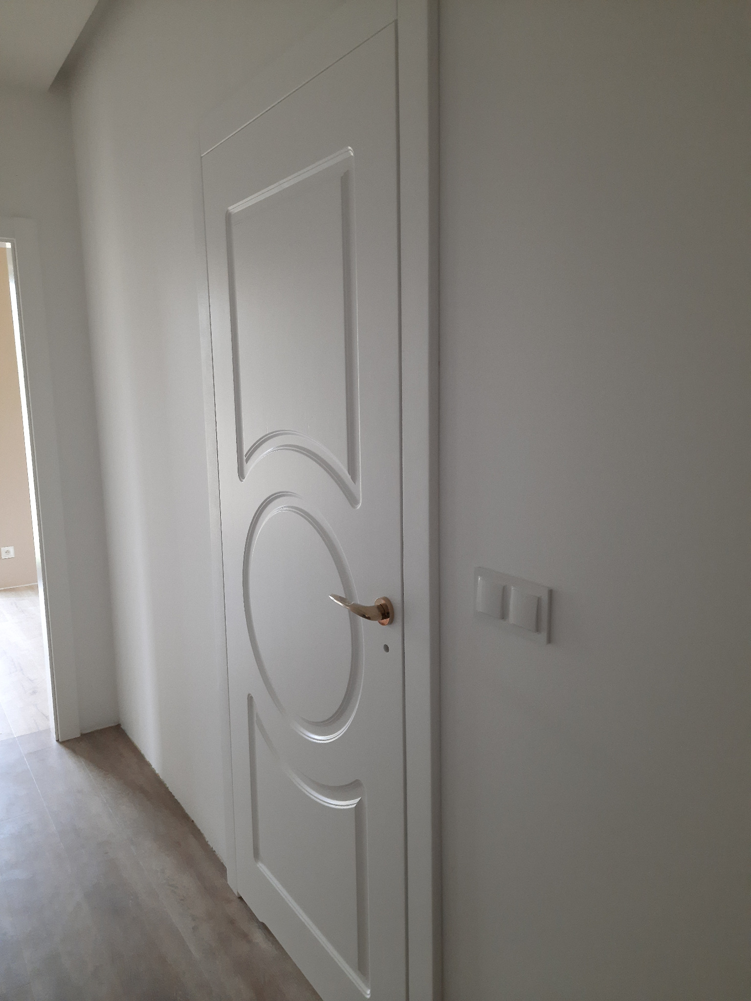 Białe drzwi wewnętrzne z okrągłym motywem dekoracyjnym i złotą klamką, zamontowane w jasnym pomieszczeniu z widocznym fragmentem podłogi i włącznikiem światła.