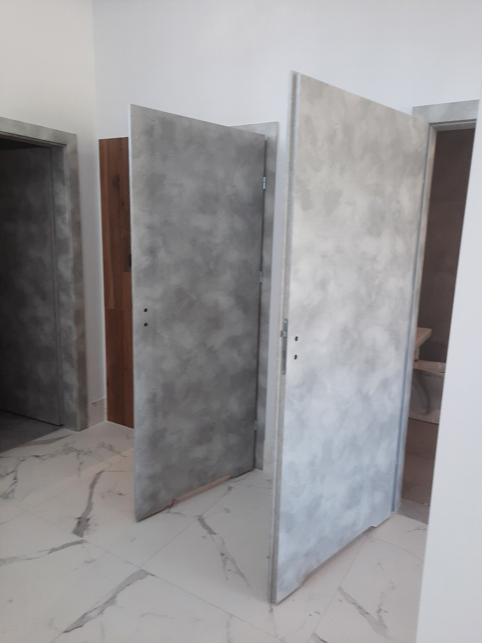 Dwa otwarte, szare drzwi wewnętrzne z efektem betonu na tle białych ścian i podłogi z marmurowym wzorem, widoczne zawiasy i klamki.