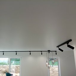 Krzysztof Kołodziejczyk - Biały sufit z zamontowanym czarnym oświetleniem szynowym i wiszącymi lampami. Widoczne okna z widokiem na zewnątrz. Minimalistyczny design wnętrza.
