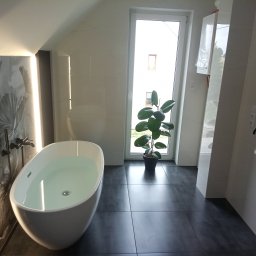 Krzysztof Kołodziejczyk - Nowoczesna łazienka w jasnych barwach z wolnostojącą wanną wypełnioną wodą, ciemną podłogą i rośliną w doniczce przy oknie. Minimalistyczny design.