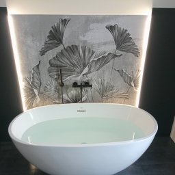 Krzysztof Kołodziejczyk - Nowoczesna, wolnostojąca wanna wypełniona wodą, na tle ściany z motywem roślinnym i podświetleniem LED. Minimalistyczna armatura w kolorze czarnym.