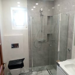 Krzysztof Kołodziejczyk - Nowoczesna łazienka po remoncie: szare płytki imitujące marmur, kabina prysznicowa ze szkła, biała umywalka i toaleta. Widoczne detale wykończeniowe.