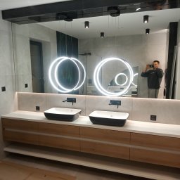 Krzysztof Kołodziejczyk - Nowoczesna łazienka z dwoma umywalkami nablatowymi, podświetlanym lustrem i drewnianą szafką. Widoczne czarne baterie i oświetlenie sufitowe. W lustrze odbija się osoba robiąca zdjęcie.