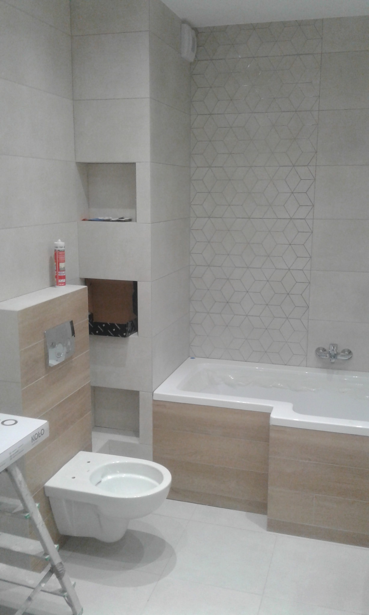 Nowoczesna łazienka w trakcie wykańczania: białe i beżowe płytki, podwieszana toaleta, wanna obudowana płytkami, wnęki w ścianie. Widoczny silikon i drabina.