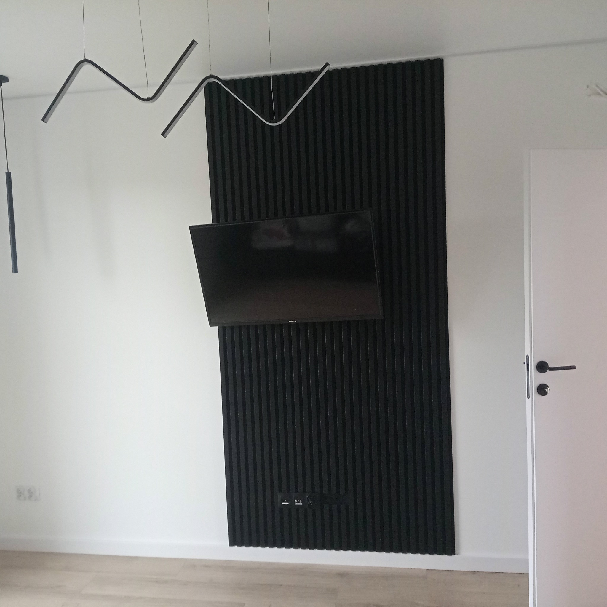 Nowoczesny salon z czarną ścianą z lameli, na której wisi telewizor. Minimalistyczne oświetlenie sufitowe i jasna podłoga. Widoczny fragment białych drzwi.