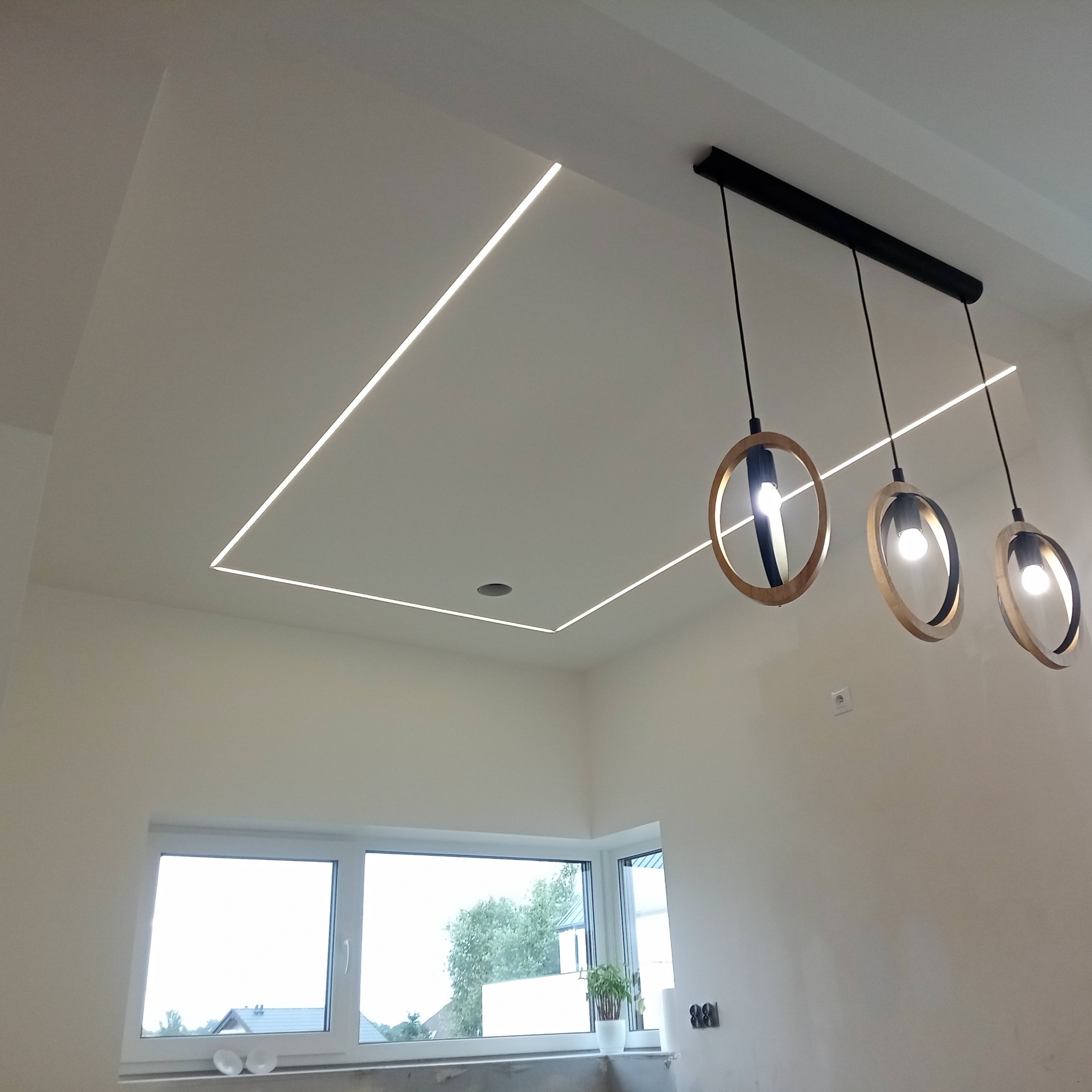 Nowoczesne oświetlenie sufitowe LED w kształcie kwadratu, uzupełnione wiszącymi lampami z drewnianymi obręczami. Widok na okno z rośliną doniczkową.