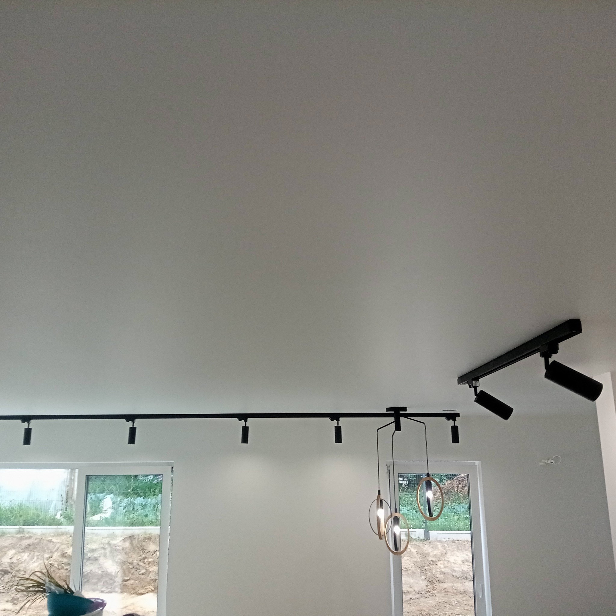 Biały sufit z zamontowanym czarnym oświetleniem szynowym i wiszącymi lampami. Widoczne okna z widokiem na zewnątrz. Minimalistyczny design wnętrza.