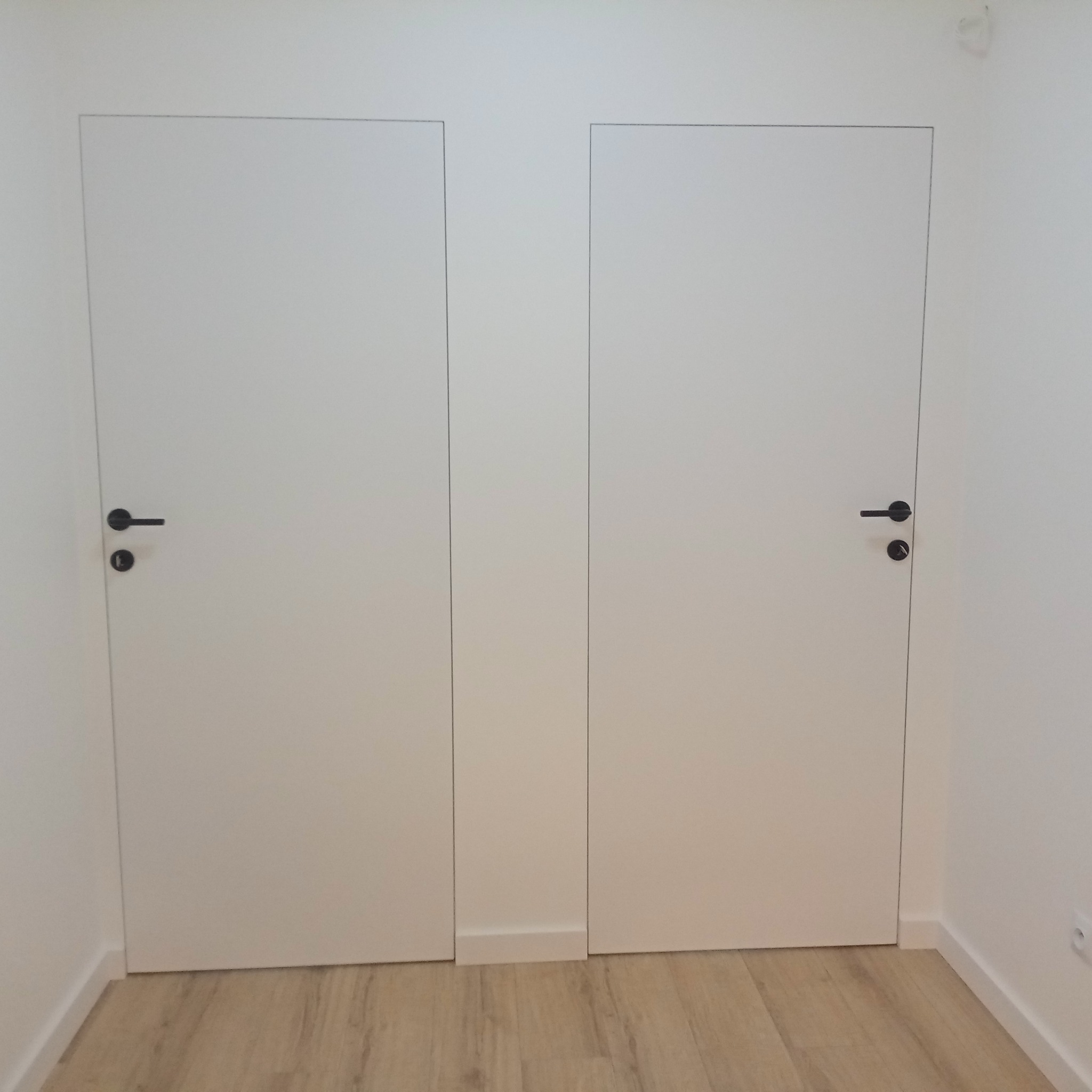 Dwa białe, minimalistyczne drzwi z czarnymi klamkami, wkomponowane w jasne ściany i podłogę z paneli. Widoczne subtelne listwy przypodłogowe.