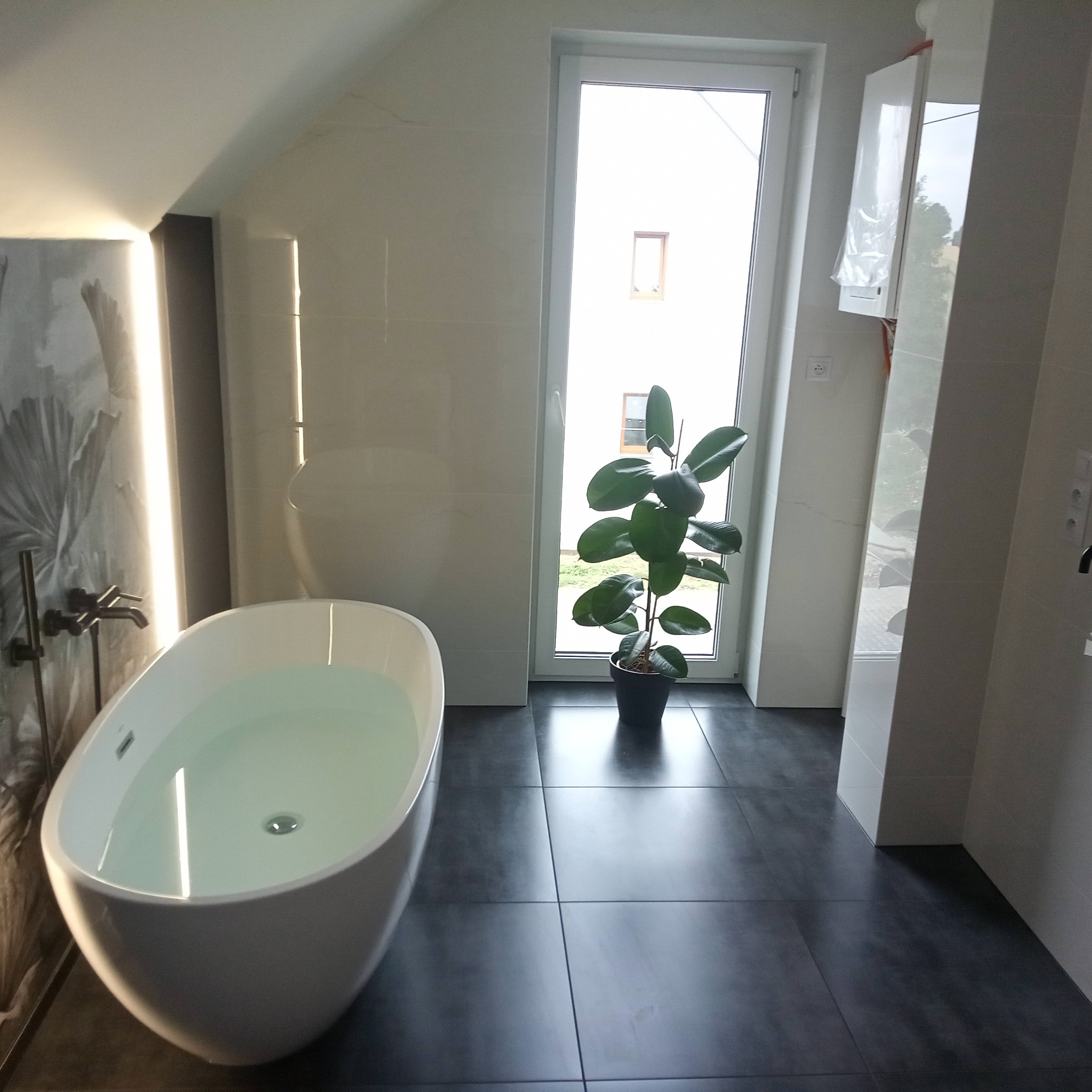 Nowoczesna łazienka w jasnych barwach z wolnostojącą wanną wypełnioną wodą, ciemną podłogą i rośliną w doniczce przy oknie. Minimalistyczny design.