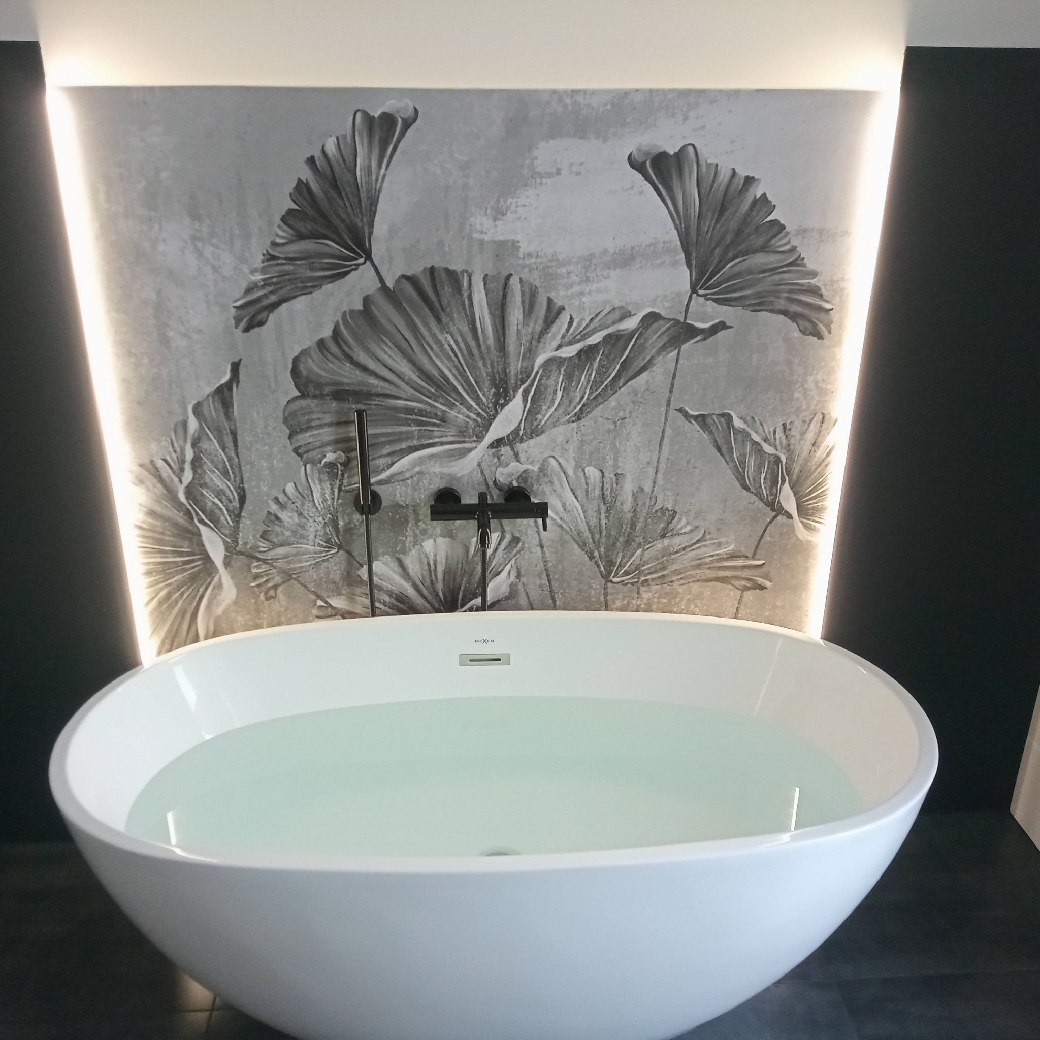 Nowoczesna, wolnostojąca wanna wypełniona wodą, na tle ściany z motywem roślinnym i podświetleniem LED. Minimalistyczna armatura w kolorze czarnym.
