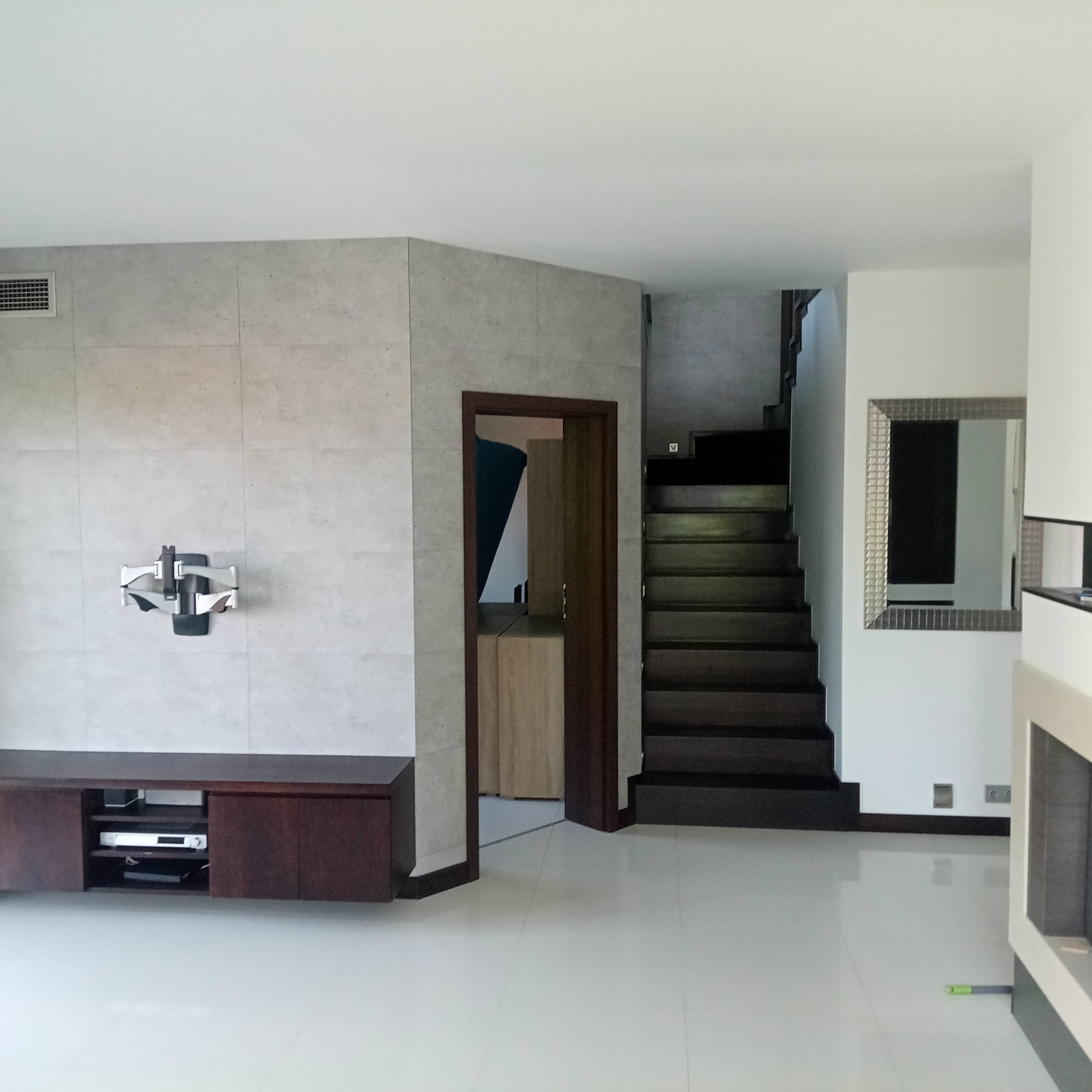 Nowoczesny salon z jasnymi płytkami podłogowymi, ścianą z płytek imitujących beton, drewnianymi schodami i szafką RTV. Minimalistyczny design wnętrza.