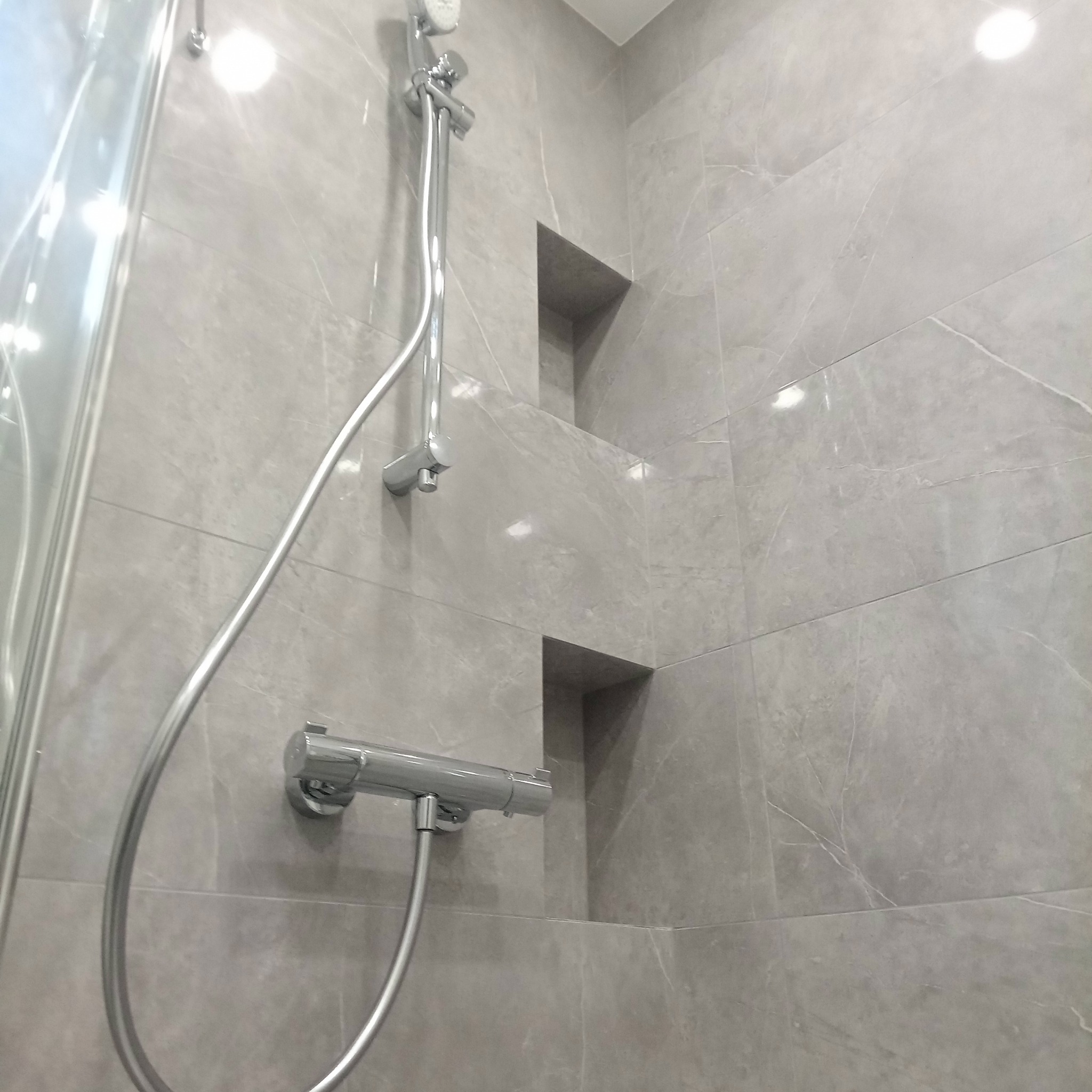 Szara łazienka z kabiną prysznicową, chromowaną armaturą i wnękami w ścianie wyłożonej płytkami imitującymi marmur. Minimalistyczny design.