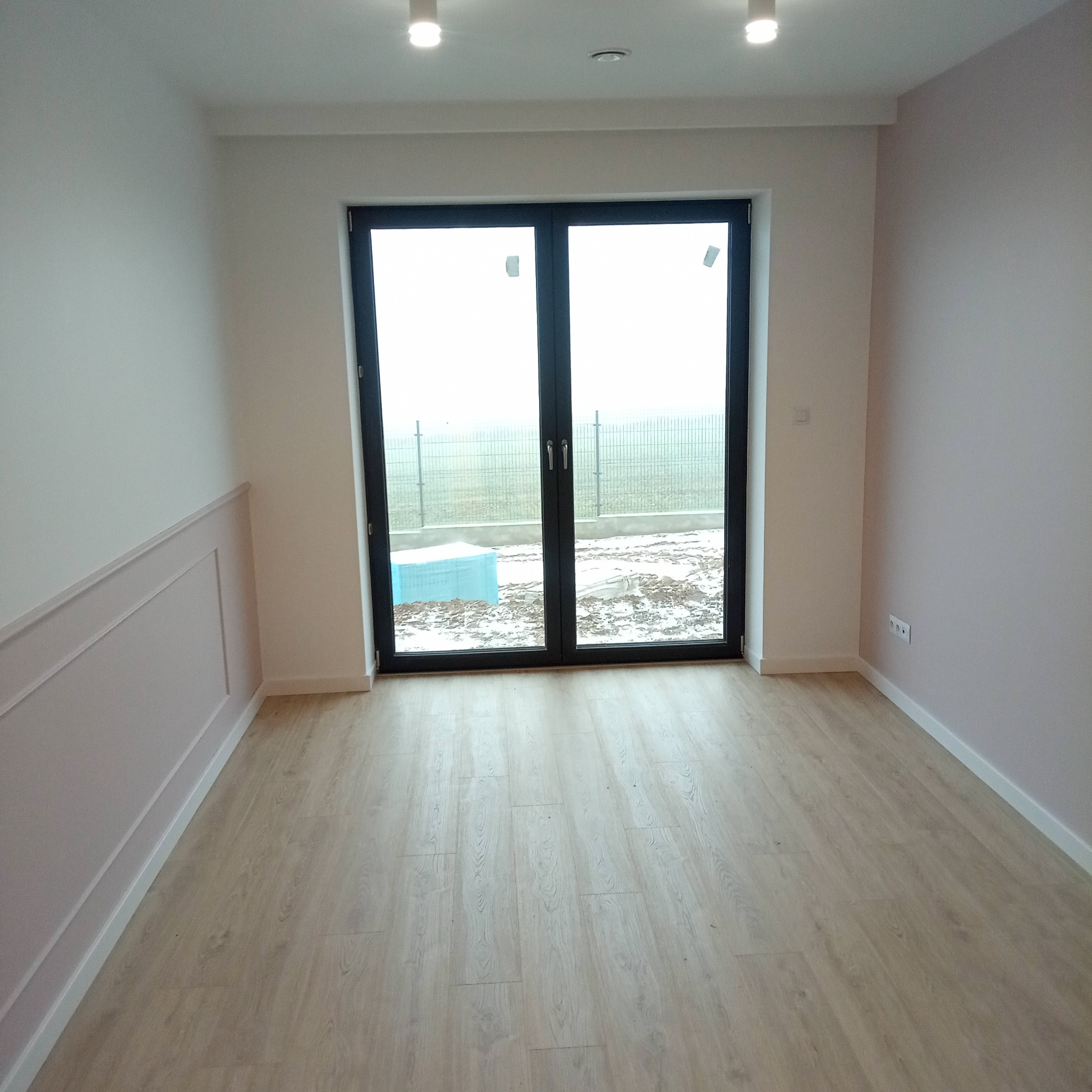 Jasne panele podłogowe w pustym pokoju z dekoracyjną listwą ścienną i dużym oknem balkonowym. Ściany w odcieniach bieli i pudrowego różu. Nowoczesne oświetlenie sufitowe.