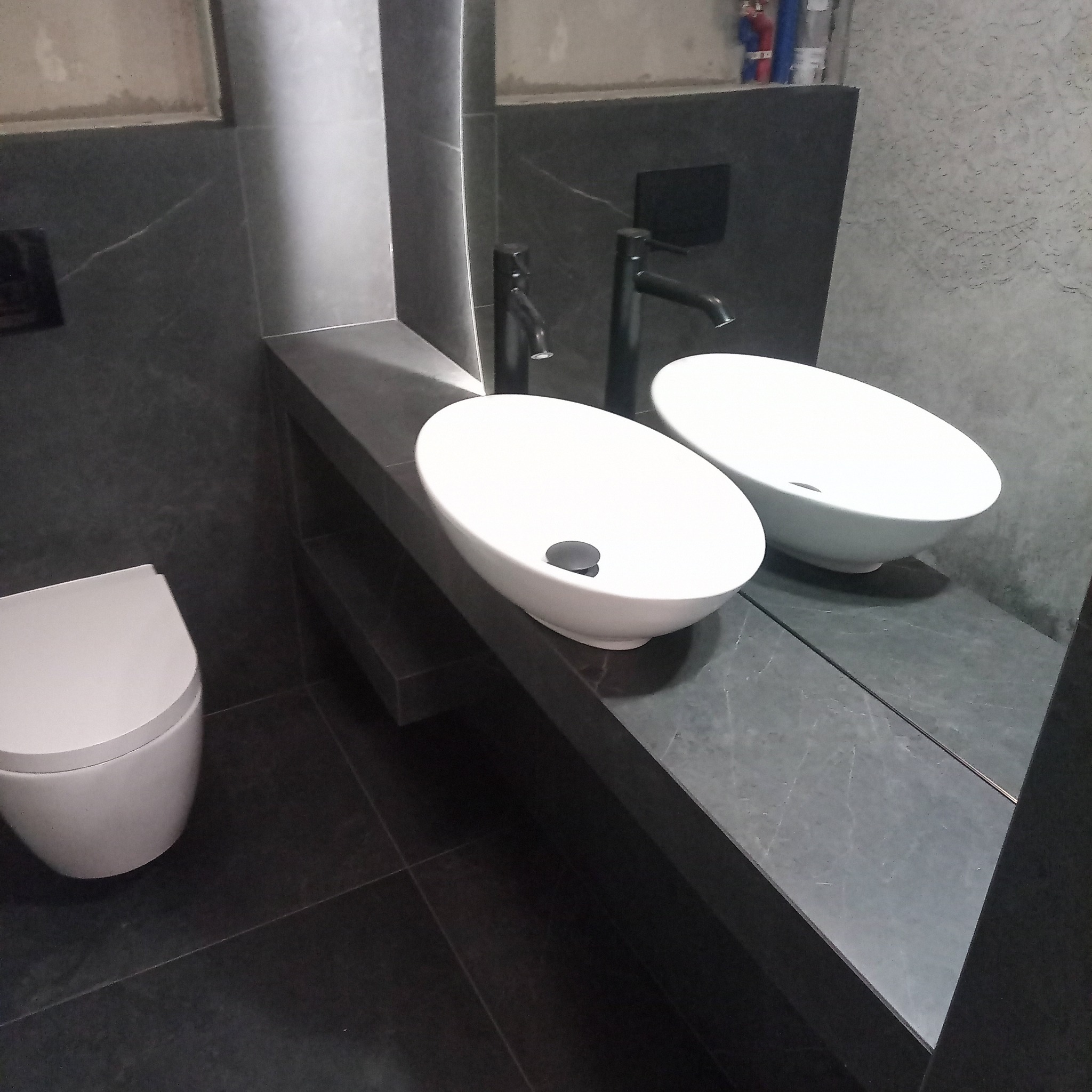 Nowoczesna łazienka z umywalkami nablatowymi i czarną armaturą na tle grafitowych płytek. Minimalistyczny design, widoczny fragment toalety.