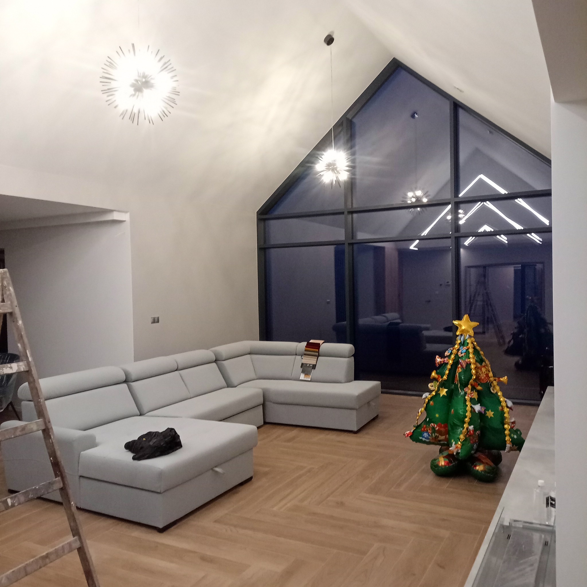 Jasne wnętrze po remoncie w Łętowni: szara sofa narożna, drewniana podłoga w jodełkę, duże okno i drabina malarska. Nowoczesny design.