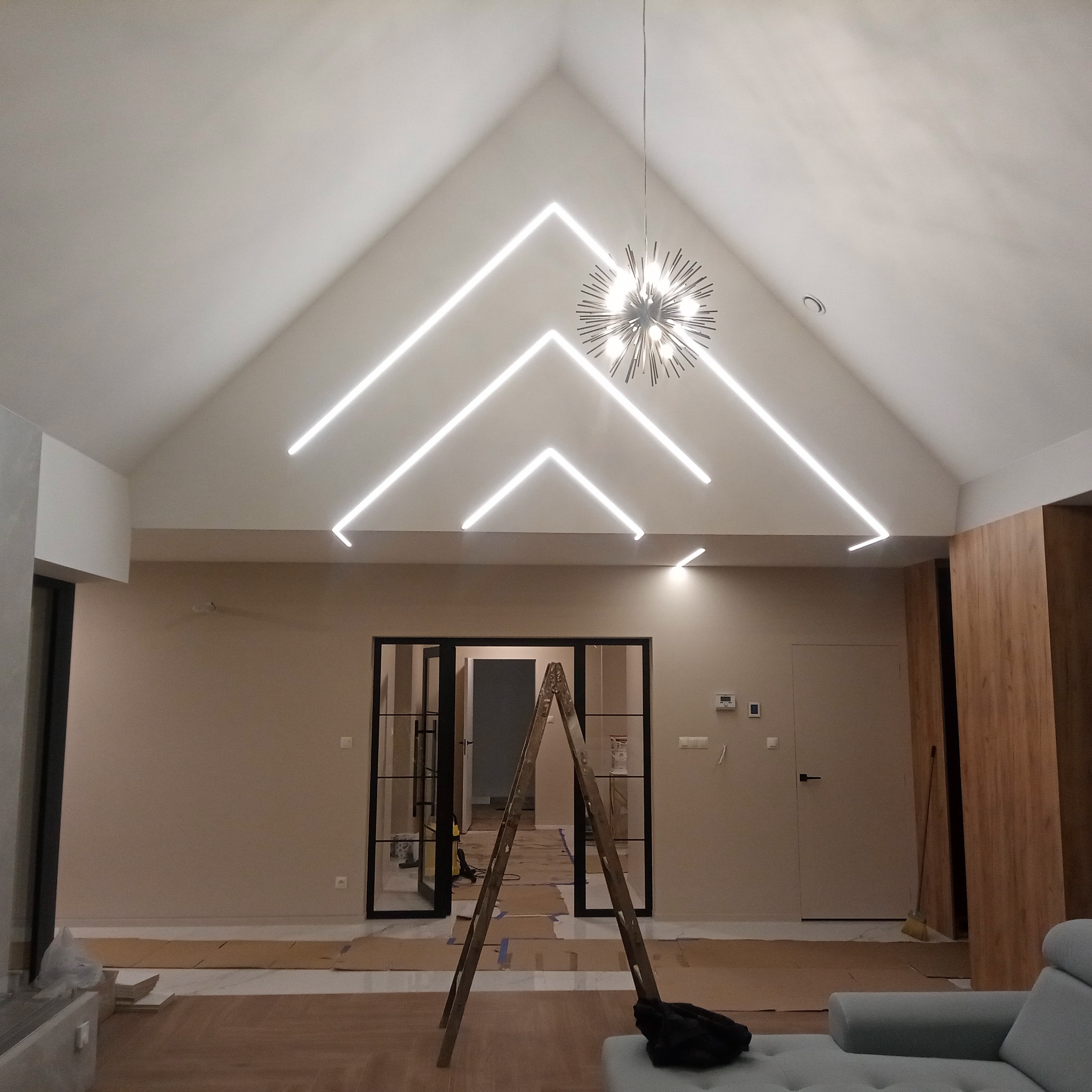 Wnętrze z wysokim sufitem i nowoczesnym oświetleniem LED w kształcie strzałek. Na podłodze kartony zabezpieczające, w tle drabina i przeszklone drzwi.
