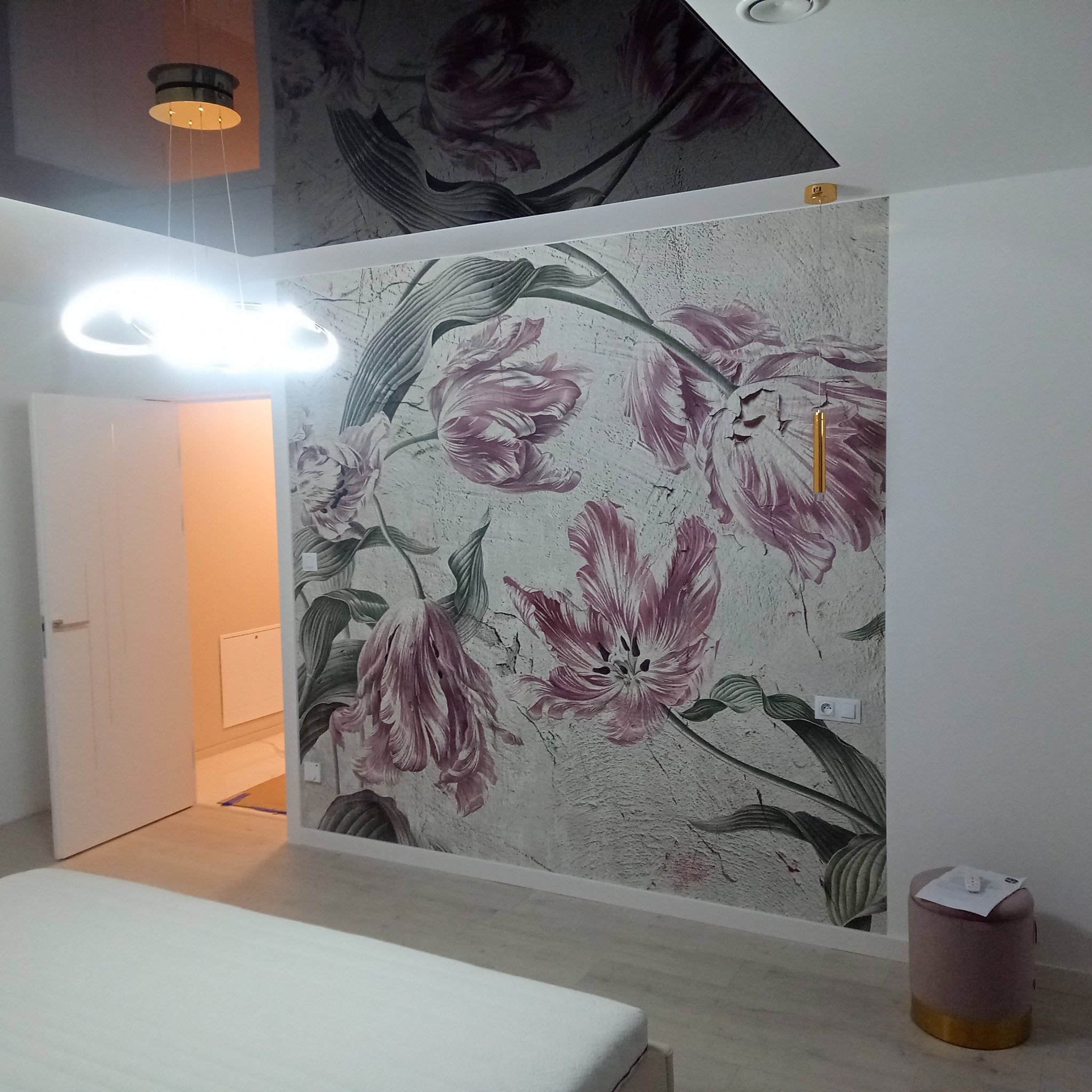 Pokój po remoncie: ściana z motywem kwiatowym, nowoczesna lampa wisząca i błyszczący sufit. Widoczny fragment łóżka i pufa. Otwarta biała futryna drzwi.