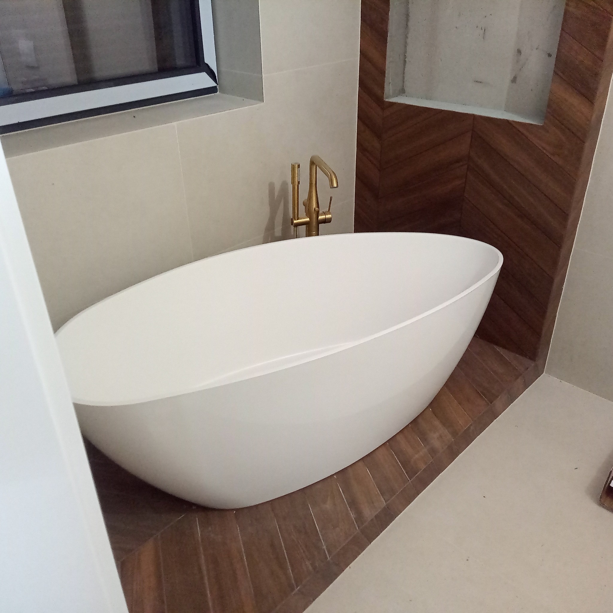 Nowoczesna, wolnostojąca wanna owalna z złotą armaturą na tle ściany z drewnopodobnych płytek ułożonych w jodełkę. Minimalistyczny design łazienki z oknem.