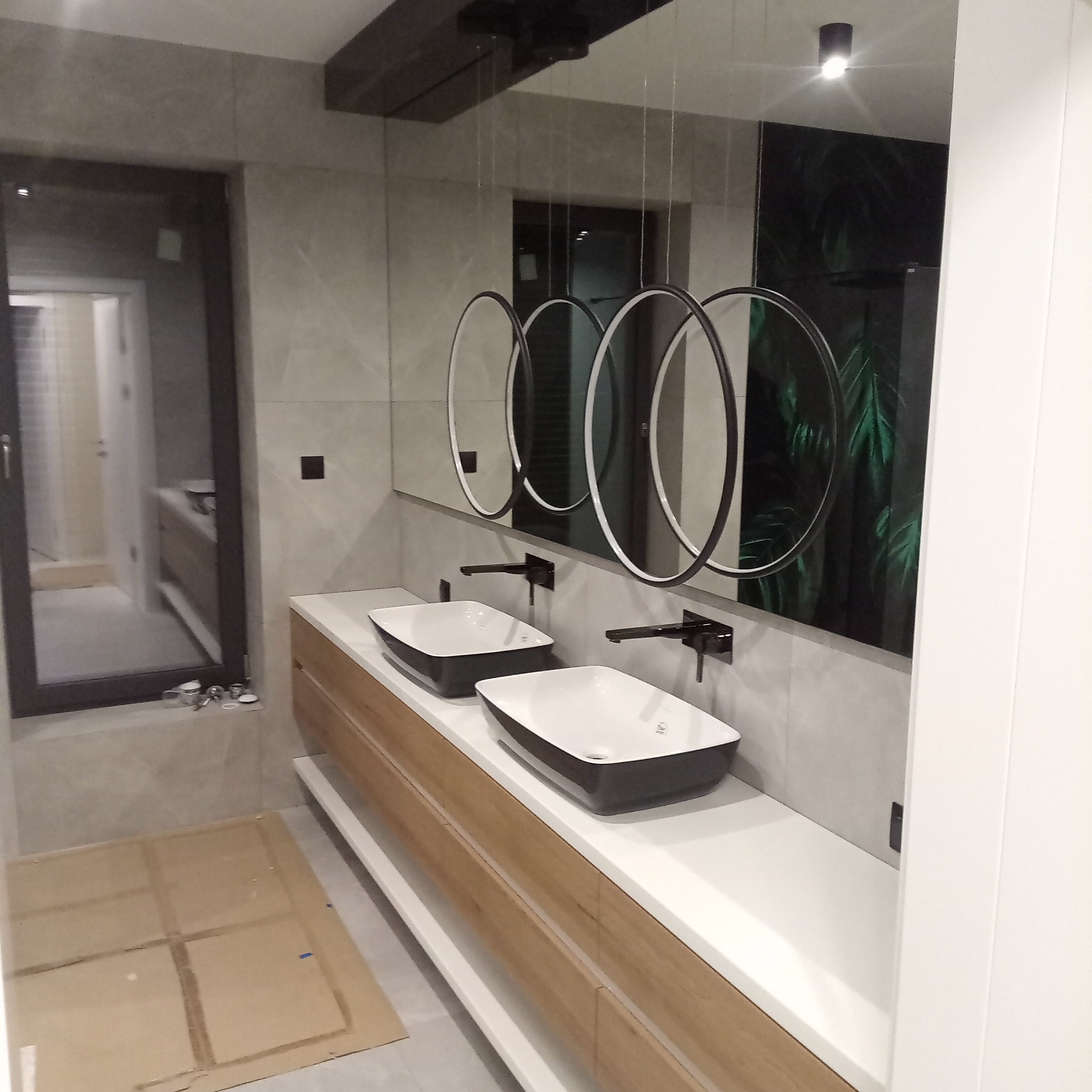 Nowoczesna łazienka z podwójną umywalką nablatową, okrągłymi lustrami z podświetleniem LED i drewnianą szafką pod umywalką. Widoczne płytki imitujące marmur i dekoracyjna tapeta.