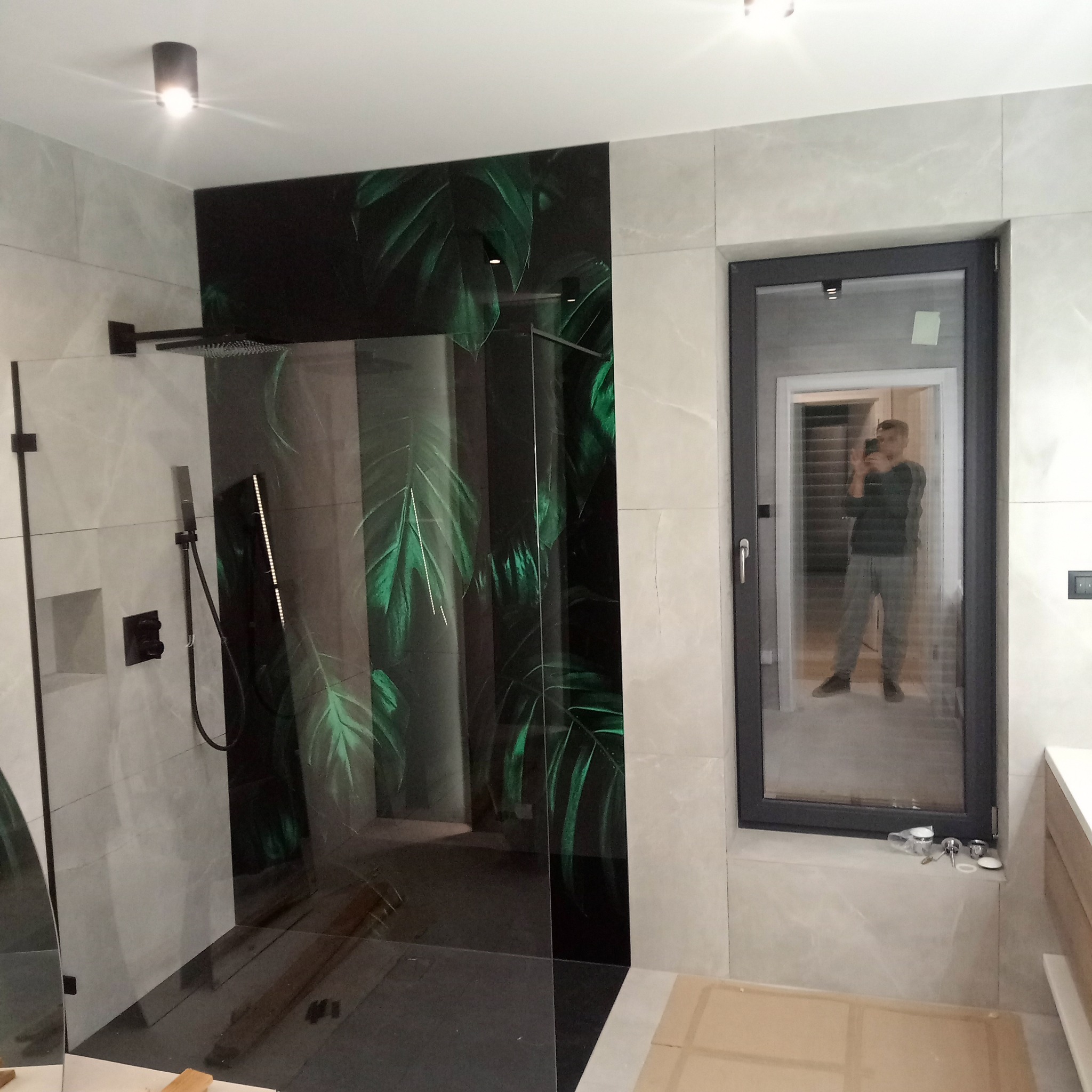 Nowoczesna kabina prysznicowa z czarnym panelem z motywem liści, szare płytki imitujące marmur, czarna armatura i okno z odbiciem osoby robiącej zdjęcie.
