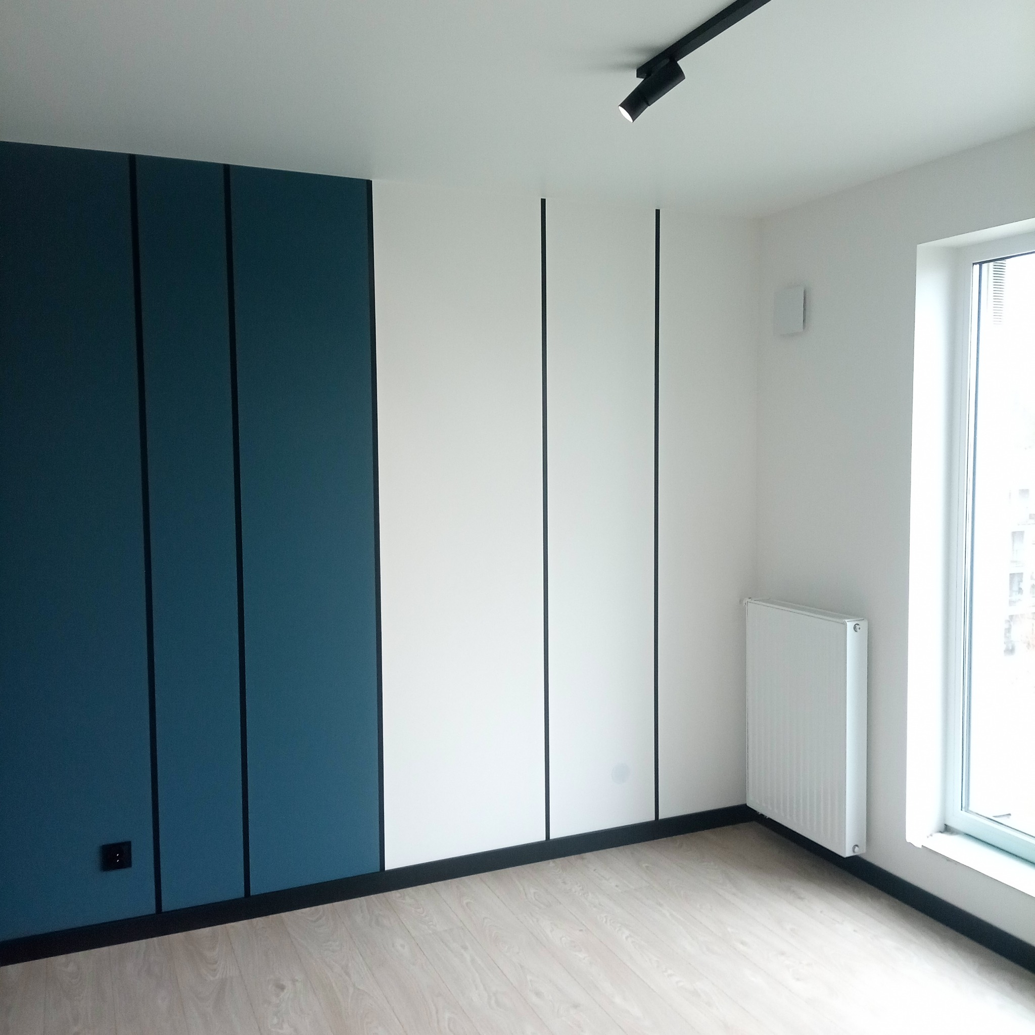 Nowoczesne wykończenie wnętrza: ściana z panelami w odcieniach bieli i granatu, czarne listwy przypodłogowe, drewniana podłoga, grzejnik i okno z widokiem na zewnątrz.