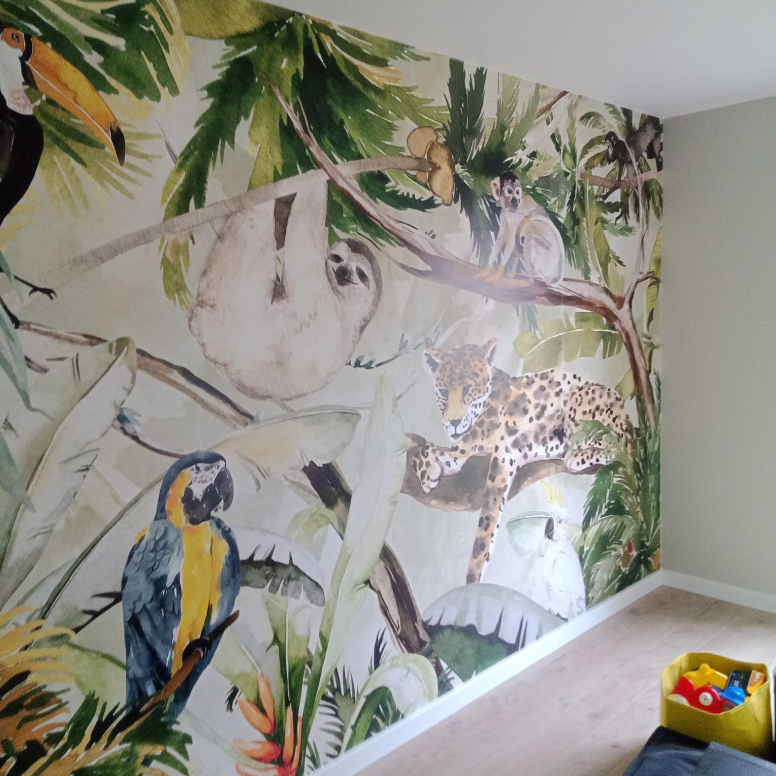 Fototapeta w pokoju dziecięcym z motywem dżungli: leniwiec, jaguar, papugi i małpy na tle tropikalnych roślin. Żółty pojemnik z zabawkami na podłodze.