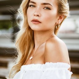 AW Fotografia Alicja Węgrzyn - Portret młodej kobiety z blond włosami, w białej bluzce z odkrytymi ramionami, z delikatnym makijażem i złotą biżuterią, na rozmytym tle.
