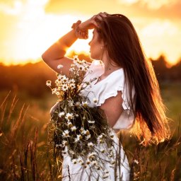 AW Fotografia Alicja Węgrzyn - Kobieta w białej sukience, trzymająca bukiet margaretek, stoi na łące w promieniach zachodzącego słońca, z ręką osłaniającą oczy.