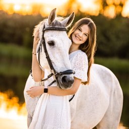 AW Fotografia Alicja Węgrzyn - Uśmiechnięta nastolatka w białej, ażurowej sukience obejmuje białego konia z czarnym ogłowiem na tle rozmytego, złotego krajobrazu o zachodzie słońca.