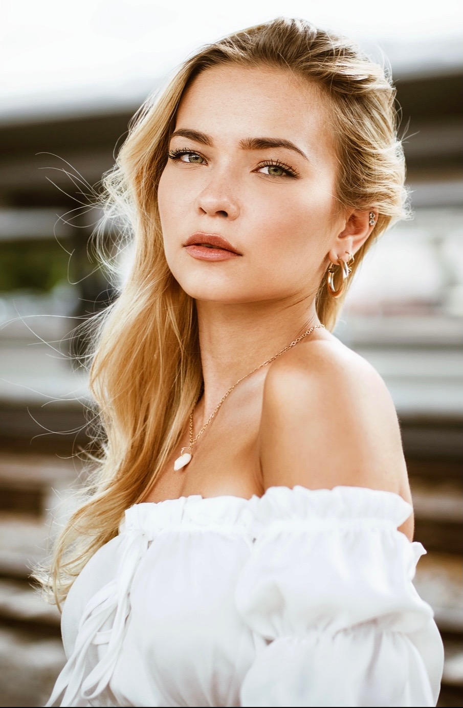 Portret młodej kobiety z blond włosami, w białej bluzce z odkrytymi ramionami, z delikatnym makijażem i złotą biżuterią, na rozmytym tle.