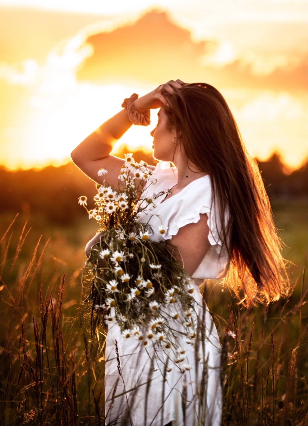 Kobieta w białej sukience, trzymająca bukiet margaretek, stoi na łące w promieniach zachodzącego słońca, z ręką osłaniającą oczy.