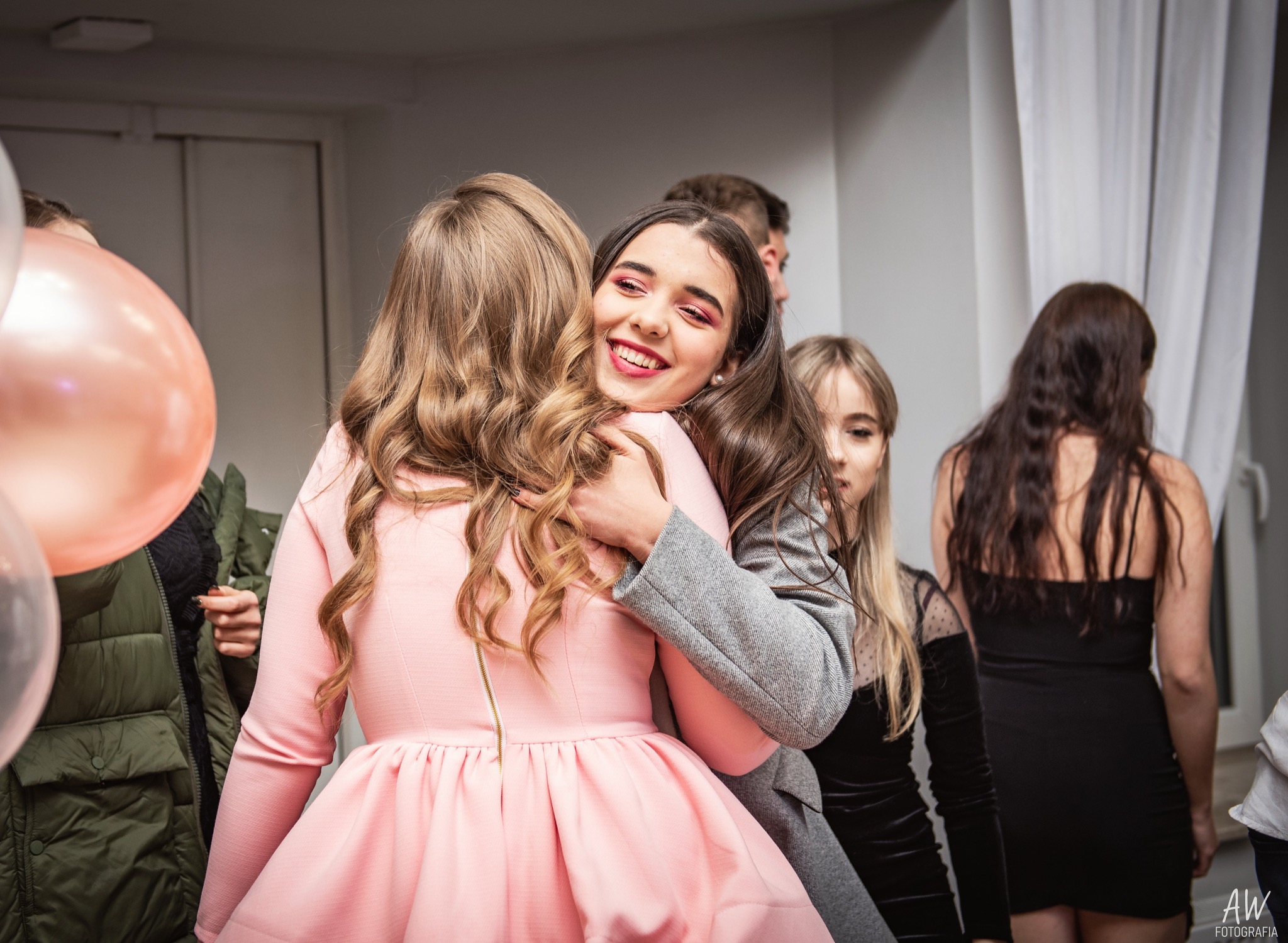 Uśmiechnięta brunetka w szarej marynarce obejmuje blondynkę w różowej sukience na tle grupy osób i pastelowych balonów.
