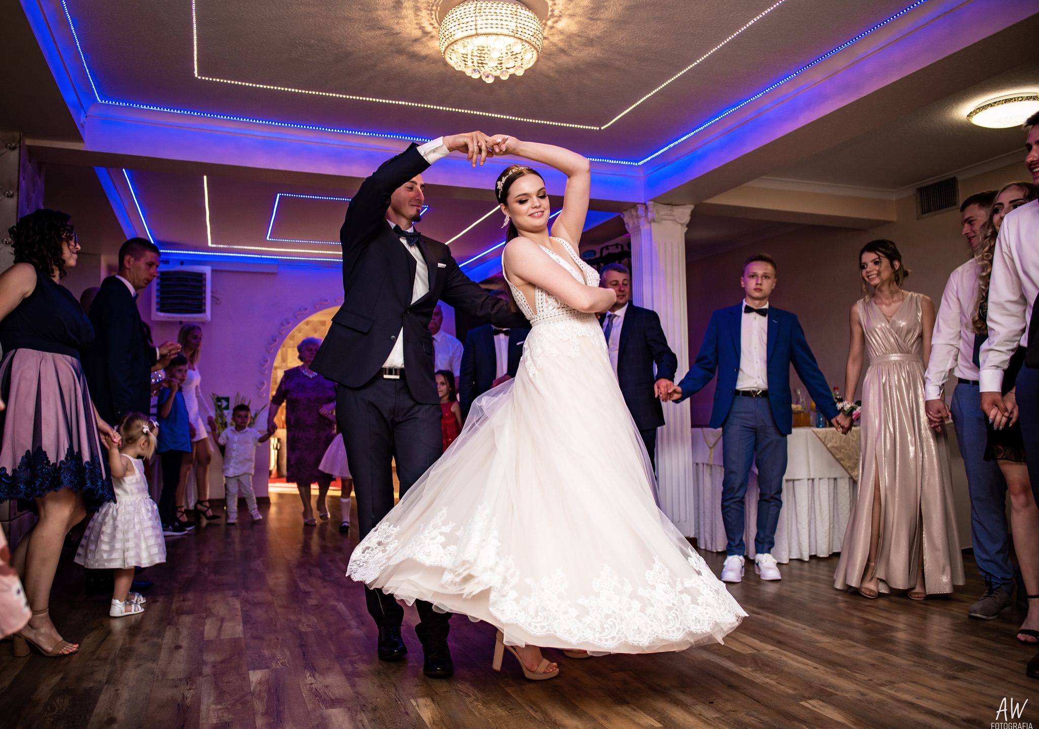 Pierwszy taniec pary młodej w sali z dekoracjami świetlnymi, goście weselni w tle.