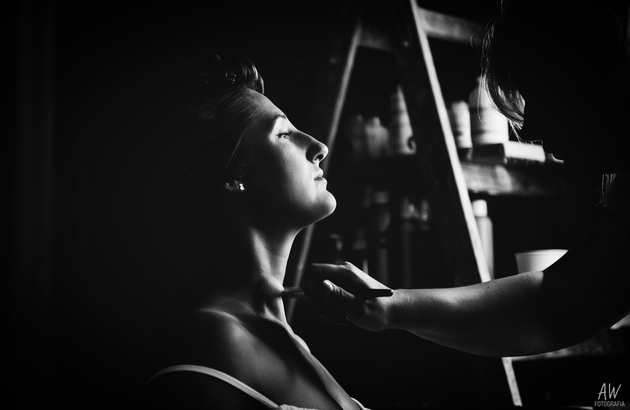 Czarno-białe zdjęcie profilu kobiety podczas makijażu szyi pędzlem, z półką kosmetyków w tle.