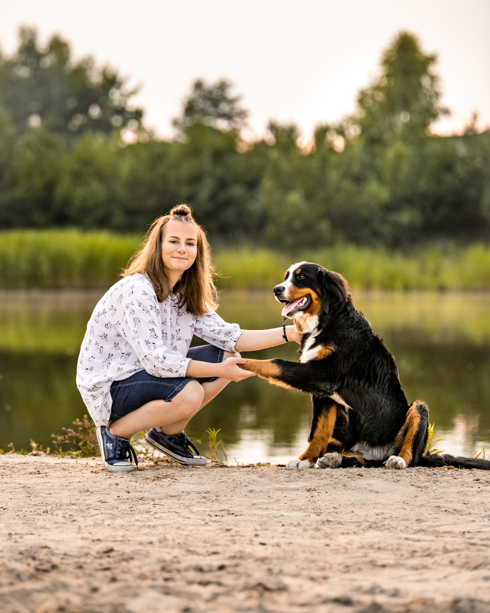 Młoda kobieta w koszuli z motywem psów kuca na piasku, trzymając łapę berneńskiego psa pasterskiego na tle jeziora i drzew.
