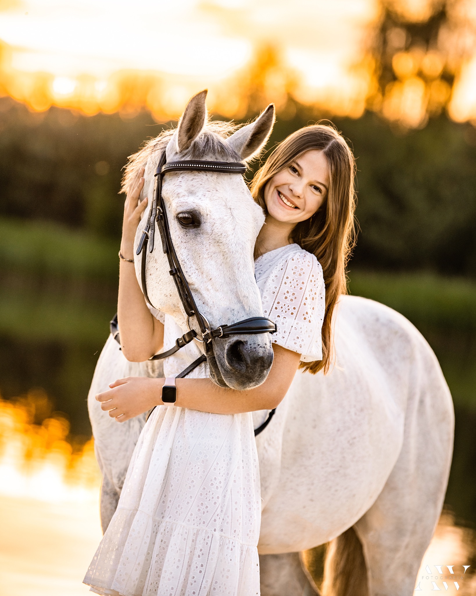 Uśmiechnięta nastolatka w białej, ażurowej sukience obejmuje białego konia z czarnym ogłowiem na tle rozmytego, złotego krajobrazu o zachodzie słońca.