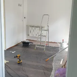 Pokój po świeżym malowaniu na biało, zabezpieczony folią malarską, widoczna drabina, kuweta z wałkiem, puszka farby i wiertarka z mieszadłem.