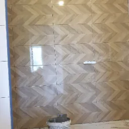 Ściana wyłożona płytkami w jodełkę, imitującymi drewno, z widocznym poziomem, wiadrami z zaprawą i narzędziami glazurniczymi na podłodze, w trakcie prac wykończeniowych.