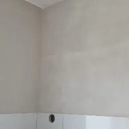 Ściana po szpachlowaniu, widoczne nierówności i przygotowanie do malowania, białe płytki w dolnej części ściany, otwór na gniazdko elektryczne.