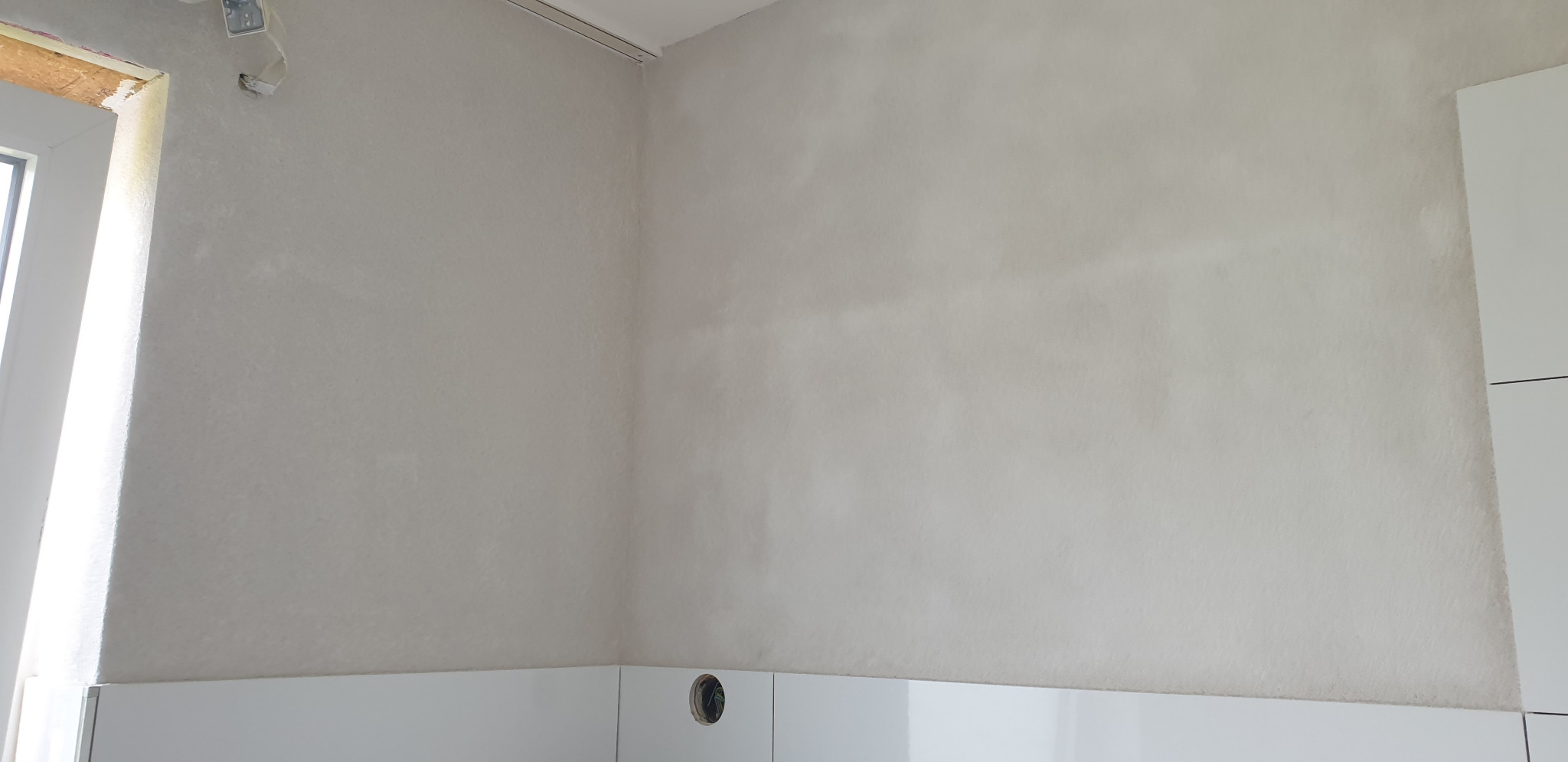 Ściana po szpachlowaniu, widoczne nierówności i przygotowanie do malowania, białe płytki w dolnej części ściany, otwór na gniazdko elektryczne.